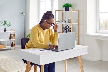 Young African American woman in glasses sitting at her desk at home, working on her modern laptop...
