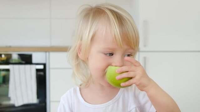 Cute blond child, girl age 2 eats half of a green apple on kitchen background, video