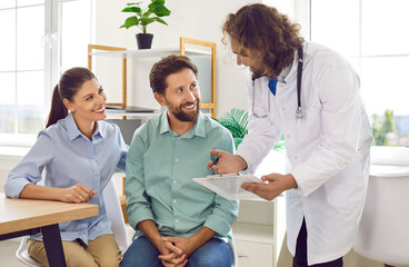 Happy young couple sitting at the desk and talking with a male family doctor therapist looking to report file with appointment during medical exam in clinic planning pregnancy. Health care concept.