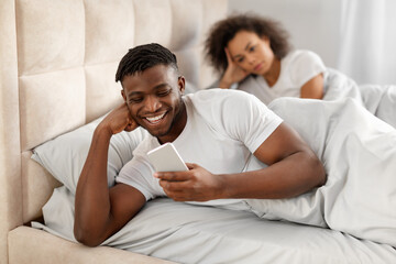 Discontented African Woman Looking At Boyfriend Texting On Phone Indoor