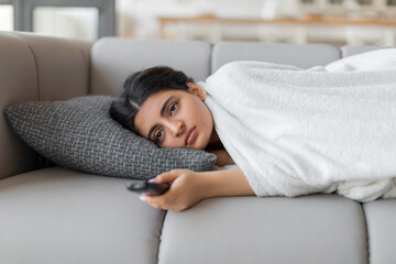 Bored indian woman with remote controller lying on sofa at home