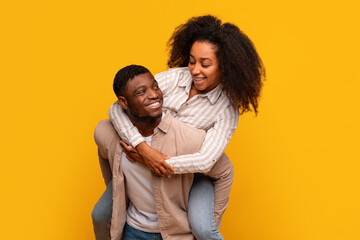 Joyful African American couple piggybacking with laughter on yellow background