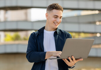 A happy young man using a laptop outdoors, dressed in a casual