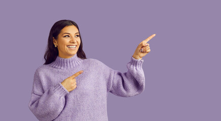 Cheerful good-humored woman in purple sweater smiling, pointing and looking to place for text. Long-haired brunet girl poses on purple background showing something interesting and exciting 