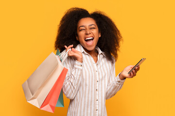 Ecstatic black female shopper with bags and smartphone on yellow - obrazy, fototapety, plakaty