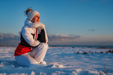 Happy young woman in winter suit sits in the snow and looking aside at sunset
