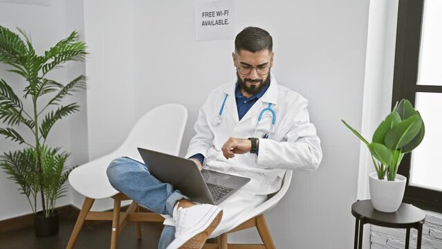 A professional young man with a beard wearing a lab coat works on a laptop in a modern indoor clinic with plants and a sign offering free wi-fi.