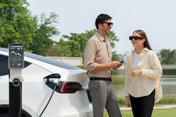 Young couple use smartphone to pay for electricity at public EV car charging station green city park. Modern environmental and sustainable urban lifestyle with EV vehicle. Expedient