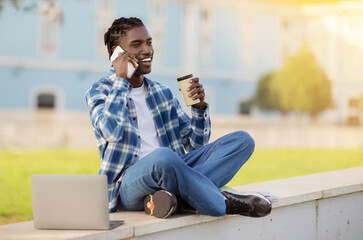 african student guy talking on cellphone and using laptop outdoors