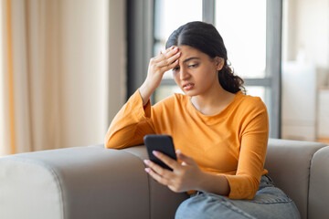 Indian woman sitting on couch at home and looking at smartphone screen