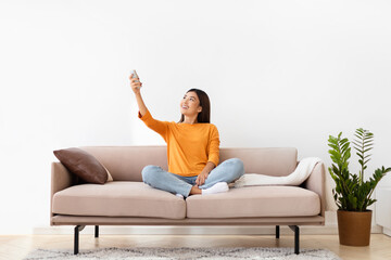 Relaxed millennial korean woman sitting on couch, turning on AC