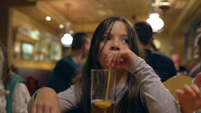 Pensive Little Girl Drinking Ice Tea with Straw at Restaurant in the Evening - Thoughtful Child Sipping Drink at Diner