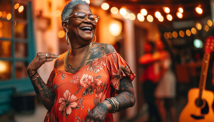 An African American senior woman with tattoos and a beautiful dress dances. Party in the evening. Background shows blurred guests, guitar, fairy lights. Copy Space.