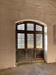 Old wooden stable door with large window in daylight, antique building. White brick wall with vintage arched doorway. Horse stable.