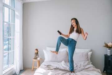 Excited woman jumping on bed with headphones and smartphone, feeling the music