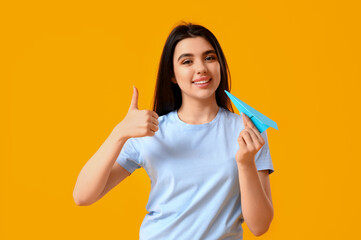 Young woman with paper plane showing thumb-up on yellow background