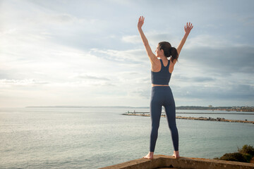 Rearview shot of a sporty woman looking out over the sea with her arms raised in celebration.