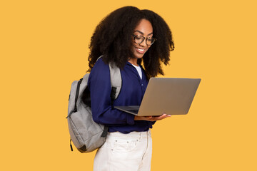 Focused black lady student with laptop and backpack on yellow background