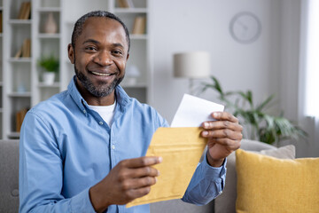 Close-up portrait of a smiling African-American man sitting at home and holding an open envelope...