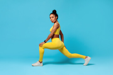 Fototapeta na wymiar African American fitness lady stretching legs lunging forward, blue background