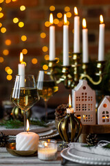 Fototapeta na wymiar Luxury elegant Christmas dinner table setting with old vintage golden chandelier, candles, fir tree branches, wooden furniture. Porcelain houses as centrepiece, garland with lights on background