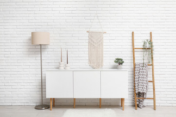 Fototapeta na wymiar Modern white chest of drawers with ladder and floor lamp near white brick wall in room