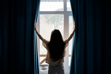 A woman with long hair stands between open curtains, welcoming natural light into a serene room through a large window, suggesting a sense of renewal or beginning.
