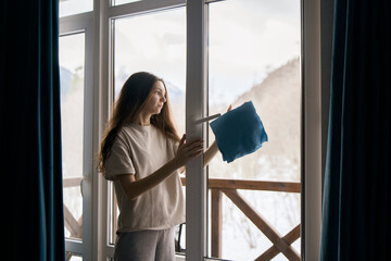 A woman meticulously cleans a windowpane using a blue cloth, with a backdrop of a tranquil,...