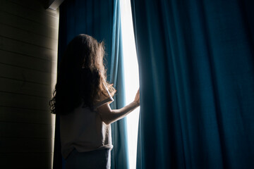 A woman with long hair stands between open curtains, welcoming natural light into a serene room...
