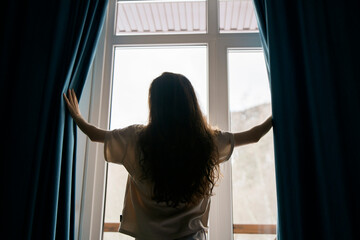 A woman with long hair stands between open curtains, welcoming natural light into a serene room through a large window, suggesting a sense of renewal or beginning.