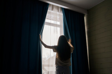 A woman with long hair stands between open curtains, welcoming natural light into a serene room...
