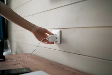 Fotobehang A persons hand is captured in the moment of inserting a two-pronged white electrical plug into a standard wall-mounted power socket, suggesting an indoor domestic or office setting. © Mihail