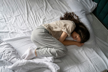 A woman is captured taking a nap, curled up in a bad wrong on a white-sheeted bed, suggesting a...