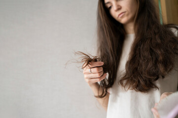 A woman appears concerned as she closely inspects the split ends of her long, wavy brunette hair,...