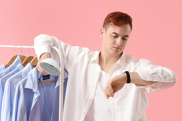 Bored young man with garment steamer checking time on pink background