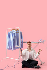 Handsome young man with garment steamer meditating near ironing board and clothes rack on pink background