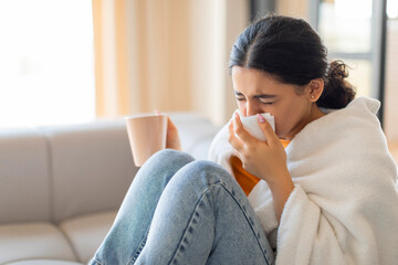 Sick young indian woman wrapped in blanket, blowing her nose with tissue