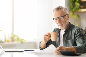 Concentrated elderly male in glasses drinking coffee while reviewing documents at home - Powered by Adobe