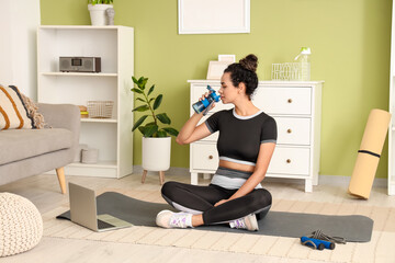 Sporty young African-American woman drinking water after training in living room