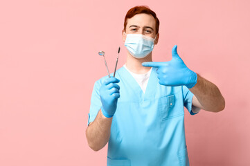 Male dentist in mask pointing at dental tools on pink background. World Dentist Day