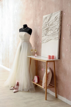 Mannequin with wedding dress, glasses of wine and engagement ring on table near beige wall. Valentine's Day celebration