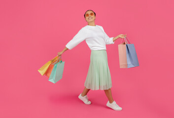 Smiling Asian woman casually walking with colorful shopping bags against pink background