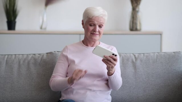 Elderly Caucasian woman sits on the couch, phone in hand, face reflecting frustration as she loses in a mobile game. The room is filled with a sense of quiet defeat as eyes fixate on the screen.