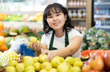 Delighted young saleswoman placing pears on food stall in large grocery store