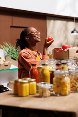 Detailed image of african american lady smelling organic farm-grown tomato in eco friendly supermarket. At the bio-food market, a youthful black woman joyfully admires newly gathered fruit. - obrazy, fototapety, plakaty