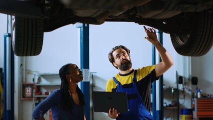 Mechanic in auto repair shop standing with customer underneath suspended car, looking together for...
