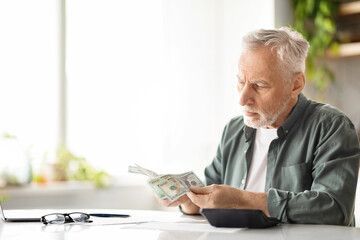 Senior male counting dollar cash while sitting at table in kitchen