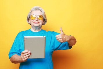 Elderly woman holding an ipad and showing thumbs up sign over yellow background