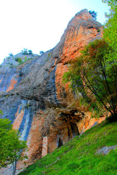 Vertical view at Grotta di Soffiano from Eremo di Soffiano near Sarnano in the Sibillini Mountains, orange and black cave in front of a brickwork wall, surrounded by stunning colored rock and trees