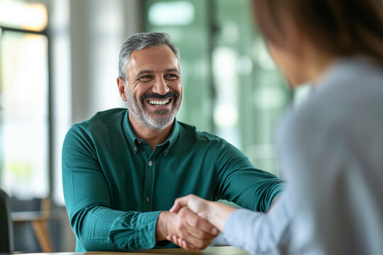 The image depicts a beaming man with gray hair, dressed in a green shirt, shaking hands with another person across a table in a bright office setting.